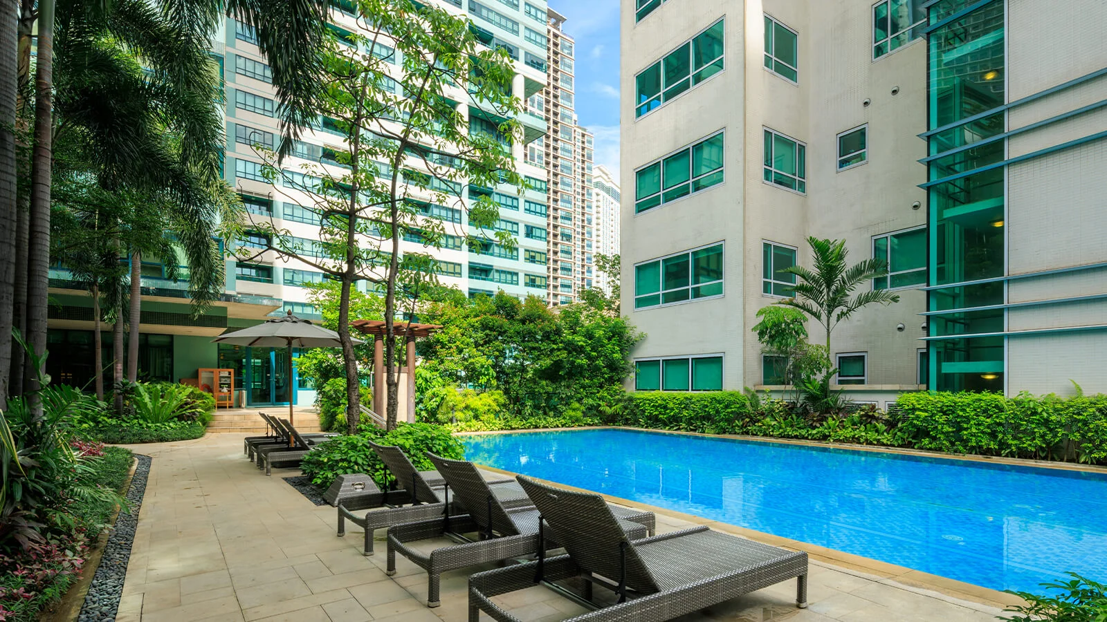 Lush greenery surrounds a sparkling pool, nestled between towering modern buildings.